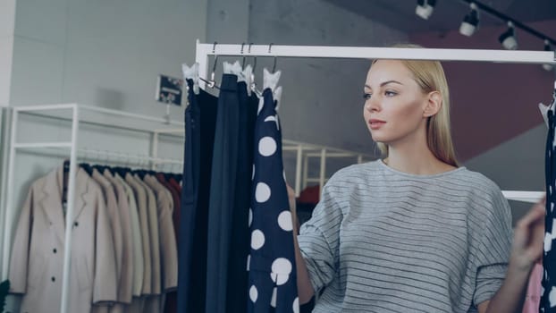 Attractive blond woman is looking at bright fashionable clothing on rails, moving them aside swiftly after checking them casually. She is enjoying shopping day and smiling.