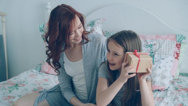 Young happy mother giving gift box to her little cute daughter celebrating birthday sitting on bed in light cozy bedroom at home