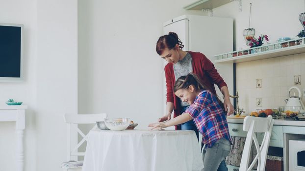 Young attractive mother teaching her little cute daughter rolling dough while cooking together in the kitchen at home on holidays. Family, food and people concept