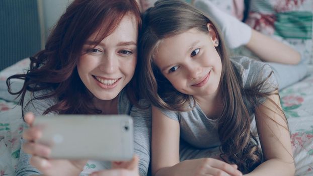 Closeup of Happy mother and little girl taking selfie photo with smartphone camera and have fun grimacing while sitting in bright bedroom at home. Family, people and technology concept