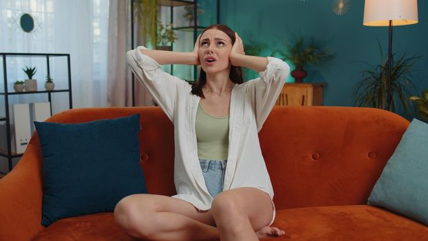 Repair work at neighbours concept. Irritated woman sit on couch cover ears with pillows annoyed by noisy neighbors suffer from headache wishes silence. Thin walls at home flat without sound insulation