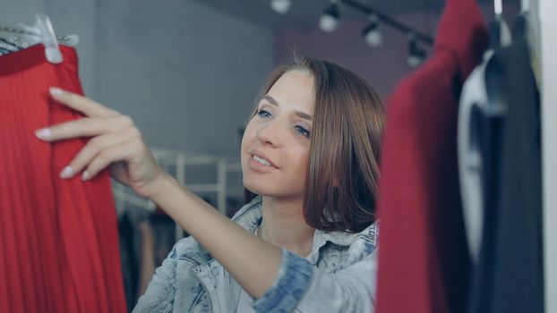 Close-up shot of attractive girl choosing clothes in boutique. She is going through fashionable tops, jumpers and skirts on hanger looking at them carefully with smile on her face.