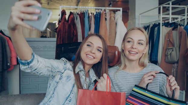 Close-up shot of cheerful attractive girls with paper bags making selfie with smartphone while sitting in clothes department in mall. First they are lauging and posing, then checking pictures