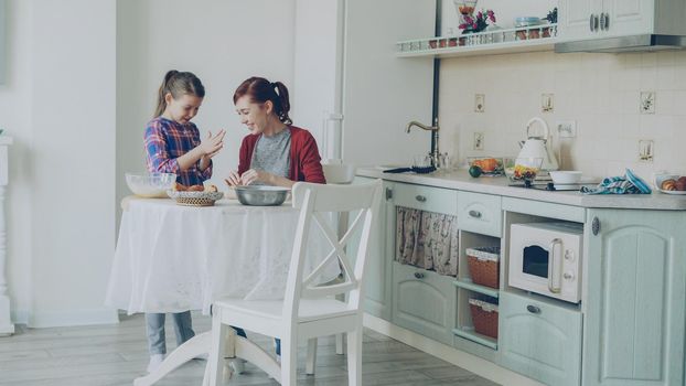 Smiling mother and cute funny daughter making cookies together using dough while sitting in modern kitchen at home. Family, food and people concept