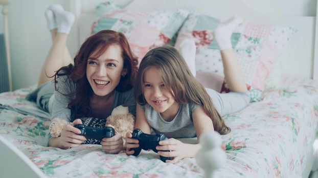 Happy young mother and cute daughter playing computer console games on TV while lying on bed at home in the morning and have fun