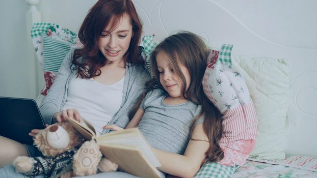 Little cute daughter with young loving mother watching funny pictures and laughing while reading fairy tale book sitting together in cozy bright bedroom at home