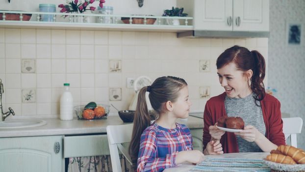 Young happy mother bringing tasty muffins to her cute daughter sitting at the table in kitchen waiting for breakfast at home