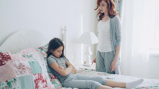 Angry mother argue scolding her upset little daughter lying on bed in cozy bright bedroom at home