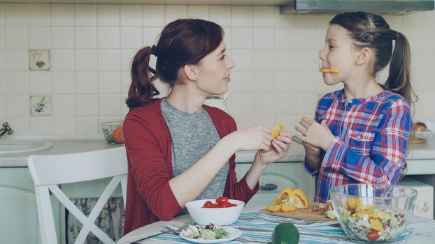 Joyful mother and cheerful daughter have fun grimacing silly with vegetables while cooking in the kitchen at home. Family, cook, and people concept