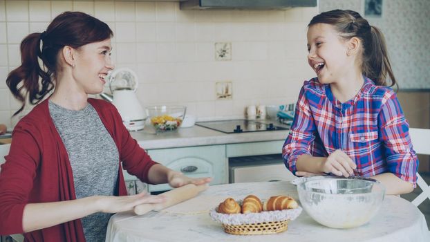 Young mother rolling dough and talking to her little cute daughter while cooking in the kitchen on weekend. Family, food, home and people concept