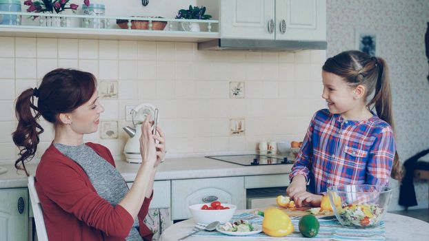 Young mother talking photo on smartphone camera of her cute daughter cooking cutting vegetables in the kitchen at home. Family, cook, and people concept