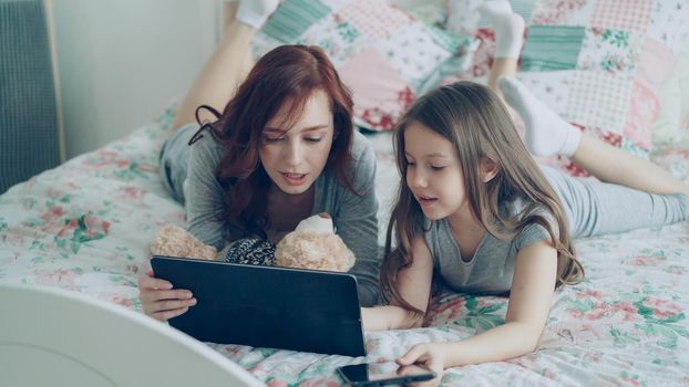 Attractive young mother and her cute daughter in pajamas laughing and looking in digital tablet while lying on bed at home in the morning