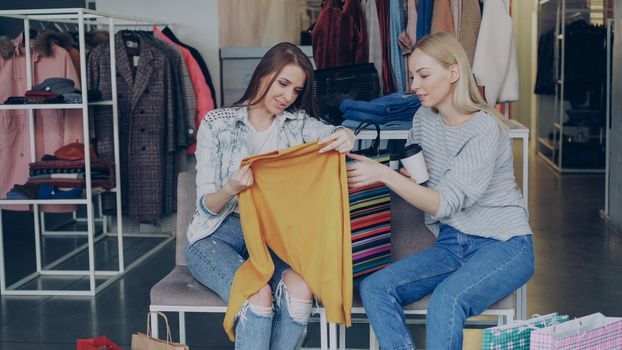 Cheerful female customer showing her friend purchased yellow jumper while sitting together in luxurious boutique. Girls are excited about price and quality of clothing.