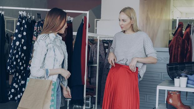 Girls are choosing clothes together while standing near rails in luxurious garments shop. They are taking skirt, fitting it to check its size and length, talking and gesturing.