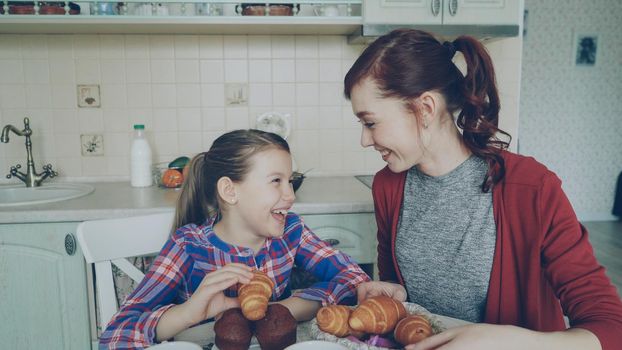 Happy young mother and cute daughter having breakfast eating muffins and talking at home in modern kitchen. Family, food, home and people concept