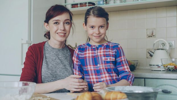 Portrait of cheerful mother and cute smiling daughter looking at camera while cooking in the kitchen on holidays. Family, food, home and people concept