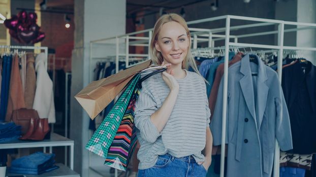 Portrait of attractive young woman standing with many colourful paper bags in modern clothing store, smiling happily and looking at camera. Stylish clothes is in background.