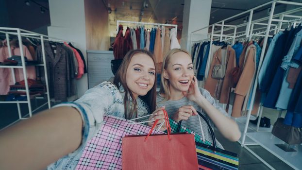 Point of view shot of attractive young ladies making selfie with paper bags in women's clothes boutique. Girls are posing, chatting, and laughing happily. Modern lifestyle concept.