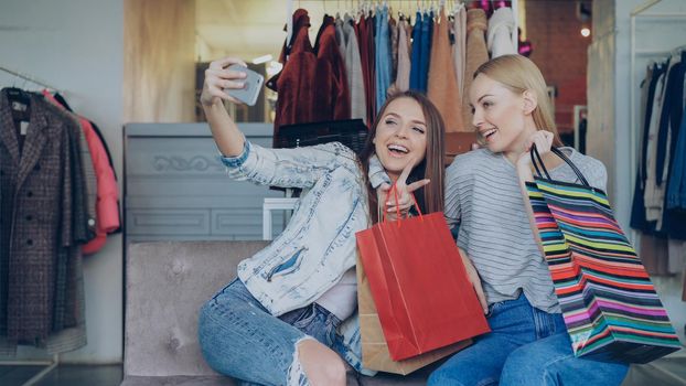 Cute girls are sitting and making selfie with colourful paper bags using smartphone in luxurious women's clothing boutique, then watching pictures together. They are smiling and laughing carelessly.