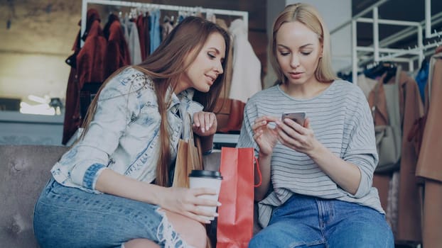 Cheerful young women chatting carelessly and using smartphone while sitting in nice clothing boutique. They are smiling and gesturing enthusiastically. Modern lifestyle concept.