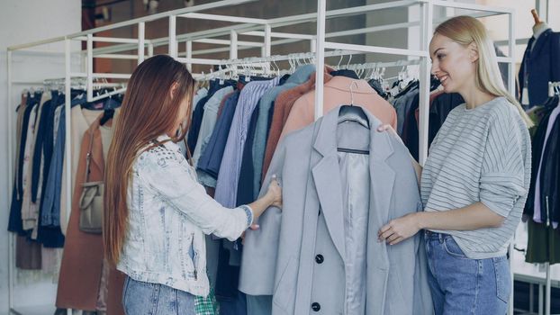 Shop assistant is helping young woman, bringing her nice coat and telling about model. Customer is touching it, comparing with other clothing on rails while chatting with saleswoman