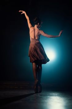 Graceful ballerina in dress dancing elements of classical or modern ballet in dark with floodlight backlight. Smoke on black background. Art concept. High quality photo