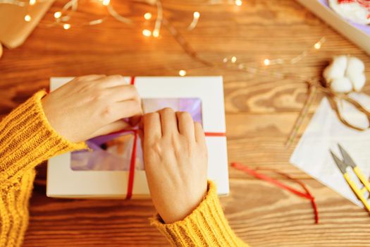 Girl's hands in knitted orange sweater tie red ribbon bow on white gift box. Garland lights, scissors and decorations in background. New Year's mood. Surprise for Birthday or Christmas.