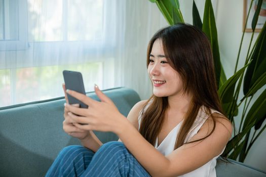 Happy Asian young woman checking social media holding smartphone at home while relaxing on sofa living room, Smile beautiful female sitting on couch using mobile phone to shopping online