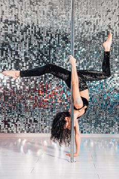 Woman doing tricks, practicing in studio with pylon. Young stripper in black dances sexually with pole in club. Poledance on shining wall background. High quality photo