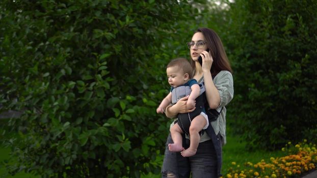 A young mother walks in the park with a child and speaks on the phone. Woman with glasses with her son. Newborn baby in a kangaroo-backpack. Close-up. 4k