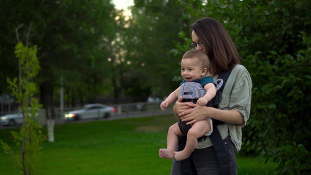 A young mother walks with a child sitting in a kangaroo backpack. Woman with glasses with a child in the park. The newborn baby laughs. Closeup. 4k