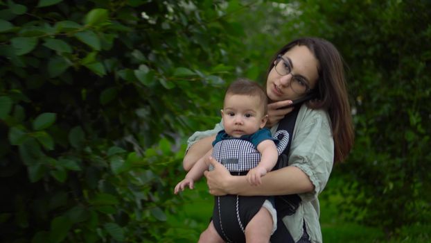 A young mother with a child walks and speaks on the phone in the park. A woman with glasses is in a hurry and clutched the phone with her shoulder. Newborn son in a kangaroo-backpack. Close-up. 4k