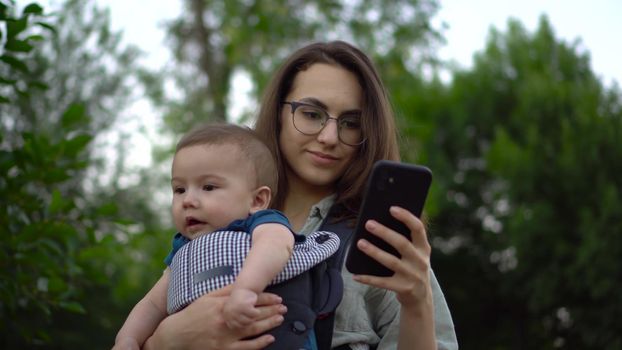 A young woman with a child stands with a phone in her hands in the park. Mom in glasses with her son in her arms. Newborn son in a kangaroo-backpack. Close-up. 4k