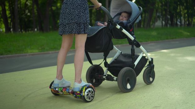 A young mother carries a stroller while standing on a hoverboard in the park. Modern walk with a child in the park. 4k