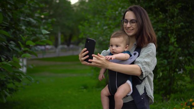 A young mother walks in the park with her little son and makes a video call on the phone. A girl in glasses walks with a newborn. A child in a kangaroo-backpack. 4k