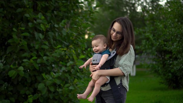 A young mother walks with a child sitting in a kangaroo backpack. Woman with glasses with a child in the park. The newborn baby laughs. Closeup. 4k