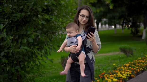 A young woman walks through the park with a child and a phone in her hands. Mom in glasses with her son in her arms. Newborn son in a kangaroo-backpack. 4k