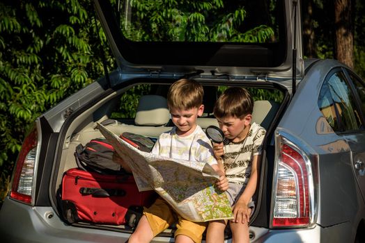 Two cute boys sitting in a car trunk before going on vacations with their parents. Kids sitting in a car examining a map. Summer break at school. Family travel by car.