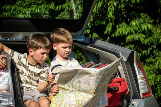 Two cute boys sitting in a car trunk before going on vacations with their parents. Kids sitting in a car examining a map. Summer break at school. Family travel by car.