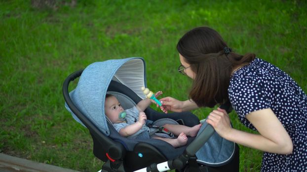 A young mother shows a rattle toy to a child. The baby lies in a stroller in the park. A woman with glasses takes care of her son. 4k