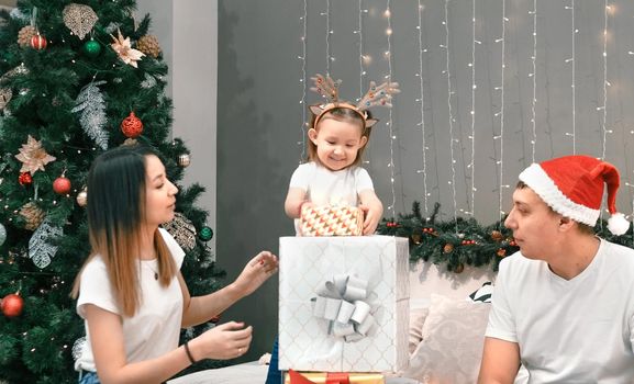 Cozy family christmas, father mother and child girl daughter play and have fun with gifts in bedroom, happy cheerful xmas holiday together, authentic festive lifestyle, gray interior