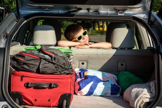 Adorable kid boy wearing sunglasses sitting in car trunk. Portrait of Happy child with open car boot while waiting for parent get ready for vocation. Family trip traveling by car concept.