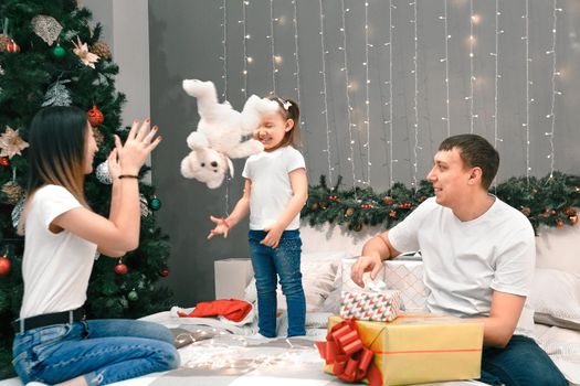 Cozy family christmas, father mother and child girl daughter play and have fun with gifts in bedroom, happy cheerful xmas holiday together, authentic festive lifestyle, gray interior