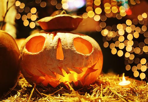 Smiling Jack O Lantern pumpkin sitting in the hay with small candles and garlands lights on background. Decoration for Halloween in club, market or cafe.