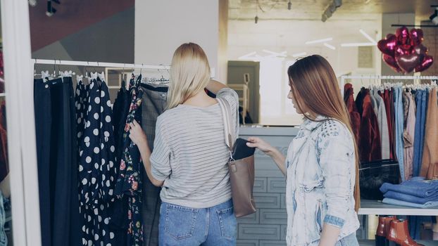 Young woman stealing wallet from female customer choosing clothes in shop. Fine boutique with clothing, bags and shoes in background