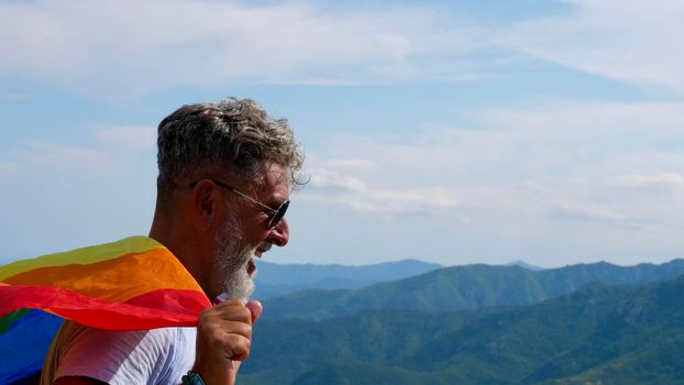 Gray-haired senior elderly Caucasian man bisexuality with a beard and sunglasses with a rainbow LGBT flag in mountains close up. Celebrates Pride Month, Rainbow Flag Day