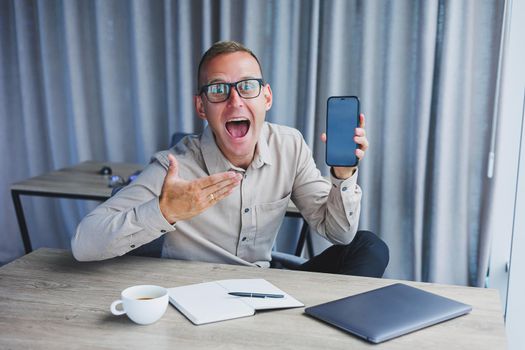 A young attractive male freelancer in glasses sits in the office and looks at the phone. Handsome blogger writing ideas on phone while sitting at wooden table in office with laptop