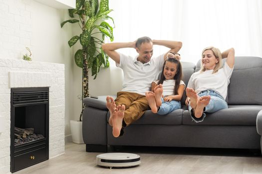 Young family resting on the couch while robotic vacuum cleaner doing its work at home. Household robots concept