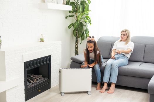 Mother and child warming hands near electric heater at home, closeup