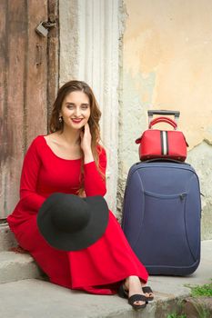 A young caucasian woman is sitting on the threshold of an old house in a red long dress with suitcase looking at the camera outdoors